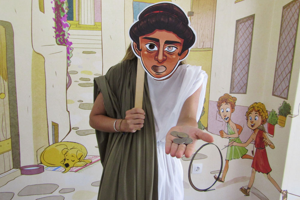 Performer in ancient Greek costume holding a character mask and replica coins at the murder mystery experience in Athens