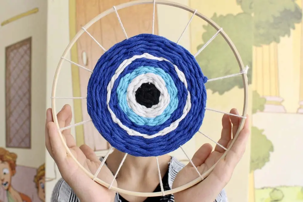 A person holds up a circular wooden loom featuring a handwoven, concentric evil eye pattern crafted from blue, white, light blue, and black yarn.