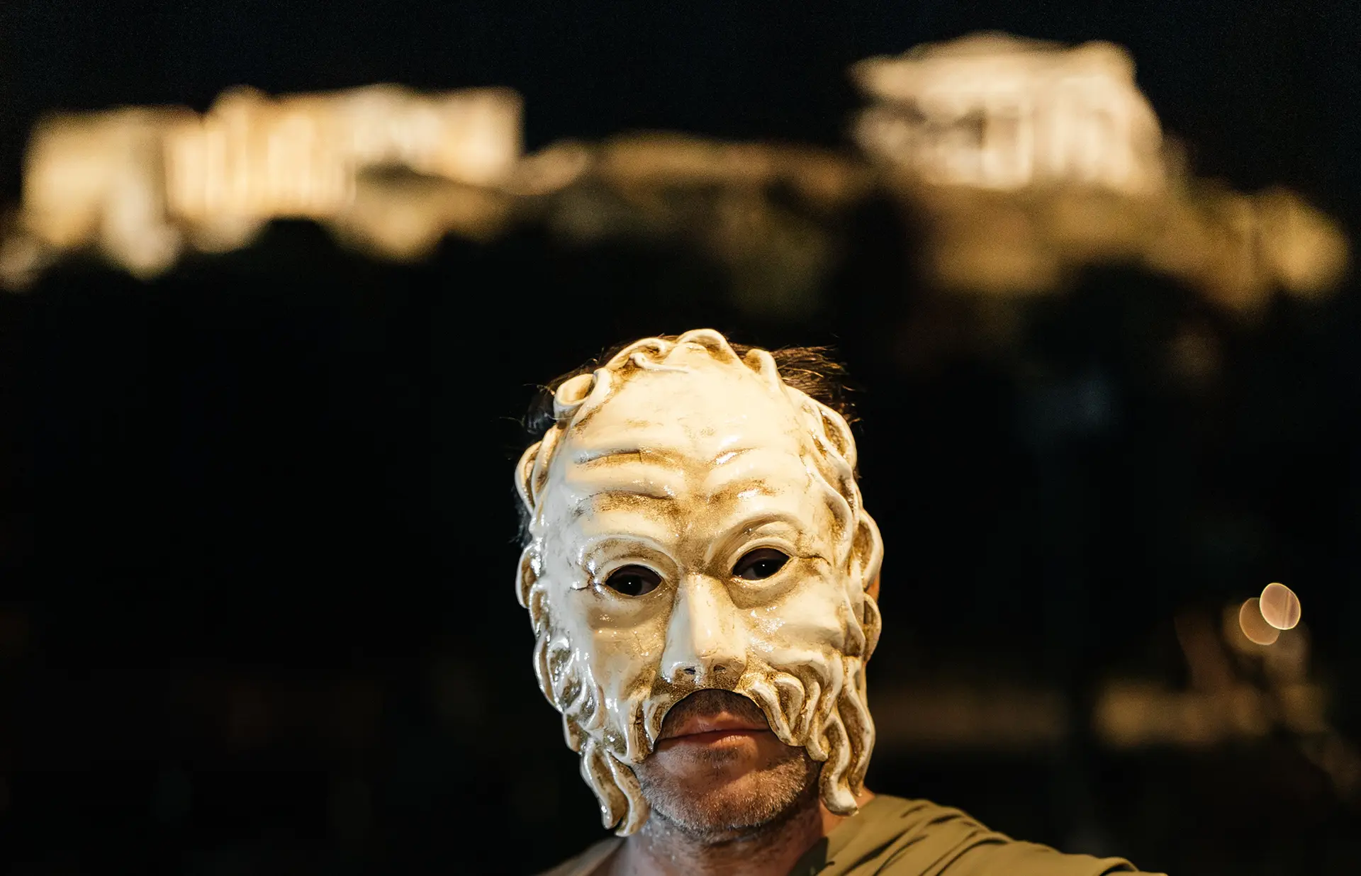 Masked performer at night with the illuminated Acropolis behind during a Theatre on the Move experience in Athens